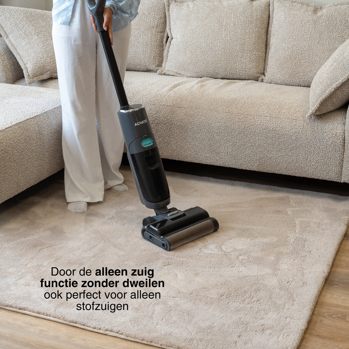 Person using a dark gray cordless stick vacuum cleaner on a light beige carpet in front of a beige fabric sofa in a living room setting