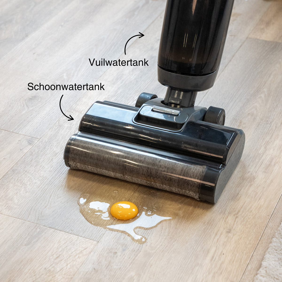 Cordless vacuum cleaner head on a hardwood floor with spilled egg yolk and white, showing labeled clean water and dirty water tanks.