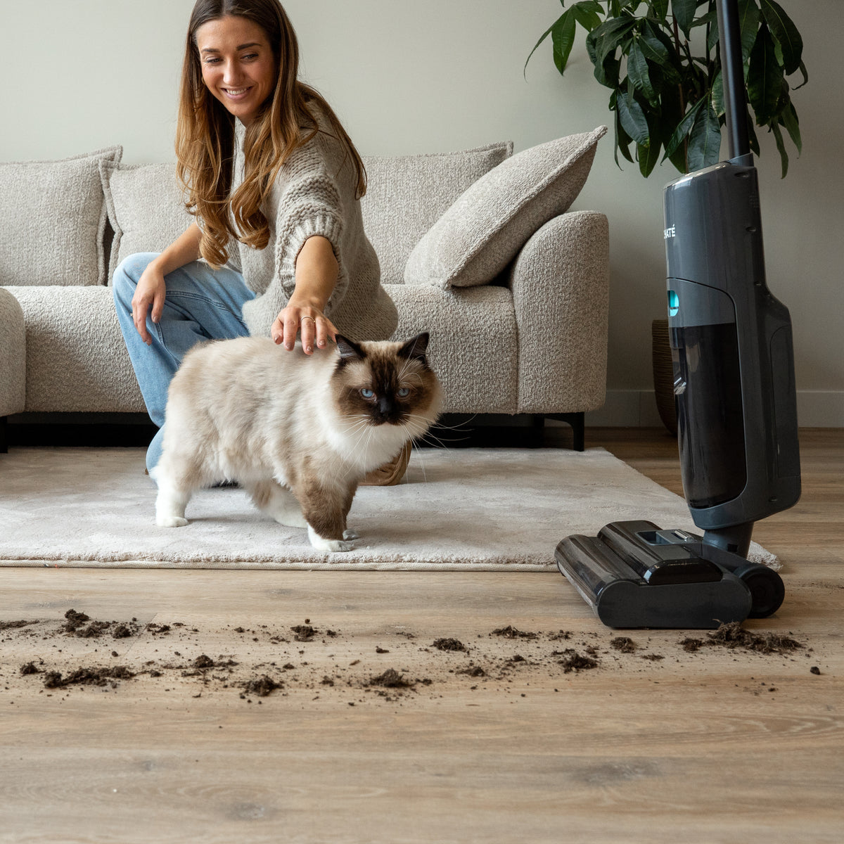 Woman petting a fluffy cat sitting on a rug next to a cordless stick vacuum cleaner cleaning dirt from a wooden floor in a living room