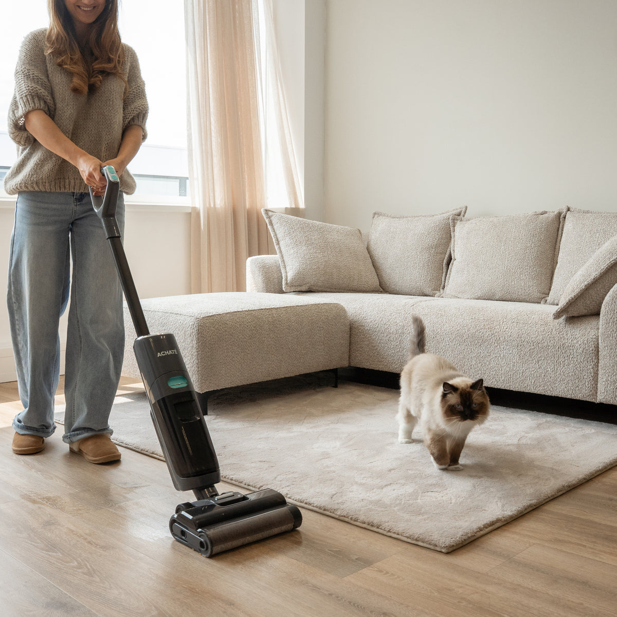 Person using a cordless stick vacuum cleaner to clean a light-colored carpet in a living room with a beige sofa and a walking cat nearby.