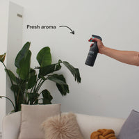 Hand spraying a black bottle near indoor green plants and a beige couch with pillows, indicating a fresh aroma in a clean living space.