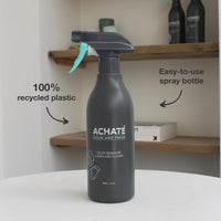 Matte black spray bottle labeled Achaté odor remover with lemon and flower scent, featuring a turquoise spray trigger on a white surface with wooden shelves in background.