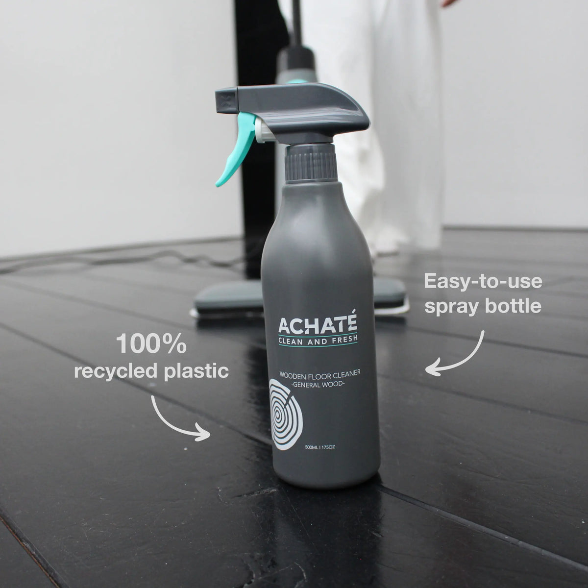 Matte gray spray bottle labeled Achaté wooden floor cleaner made from 100% recycled plastic, displayed on a dark wooden floor with cleaning device in background.