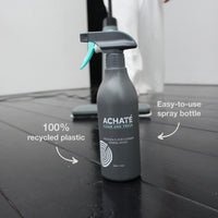 Matte gray spray bottle labeled Achaté wooden floor cleaner made from 100% recycled plastic, displayed on a dark wooden floor with cleaning device in background.