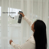 Woman using a black handheld steam cleaner on light-colored curtains near a window, demonstrating chemical-free cleaning for fabric.