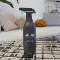 Black spray bottle labeled Achaté odor remover with lemon and flower scent standing on a reflective tiled surface in a modern living room setting.