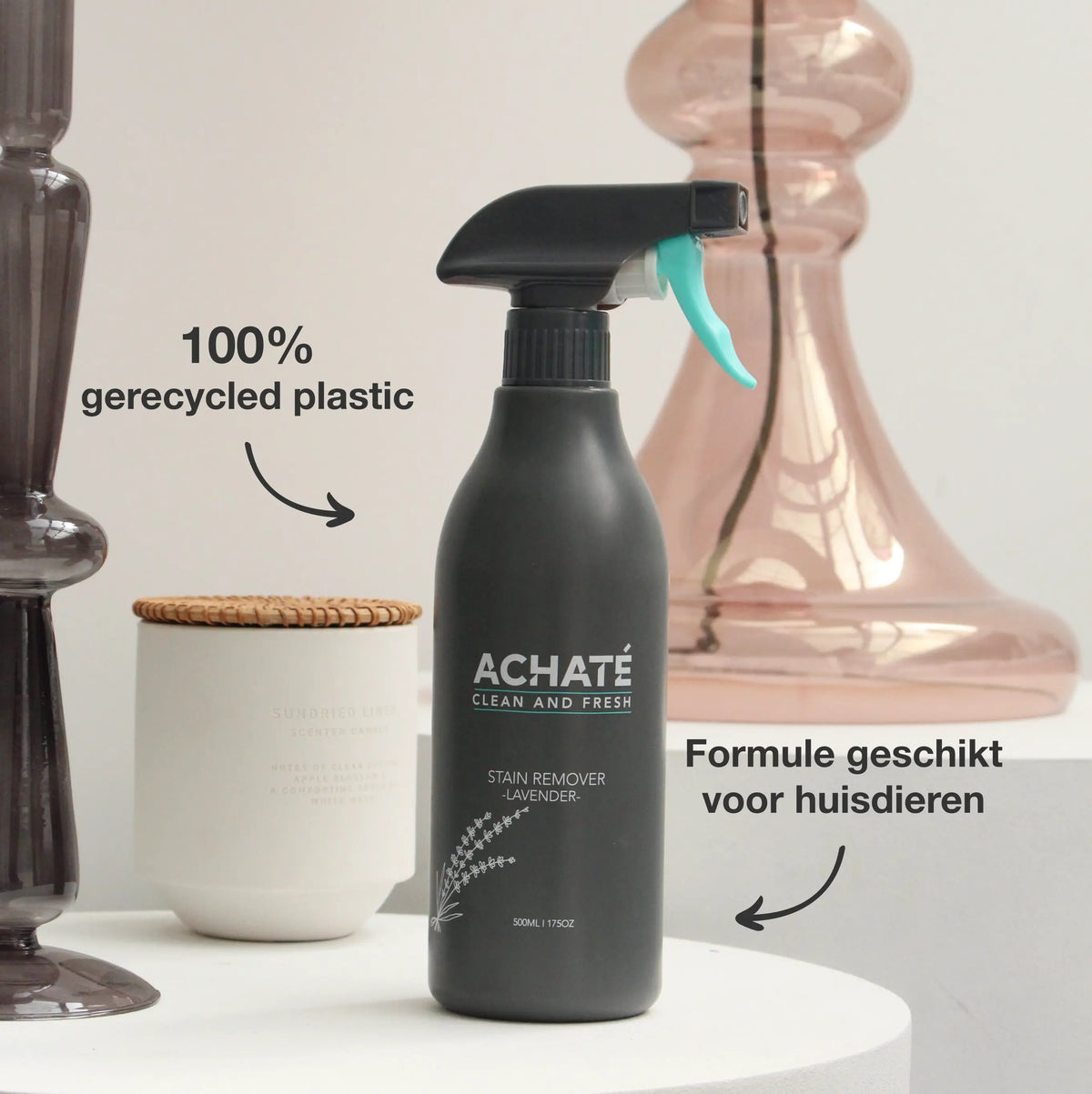 Black spray bottle of Achaté lavender stain remover with turquoise trigger, placed on white table among glass and ceramic decor pieces, labeled 100% recycled plastic and pet-safe formula.