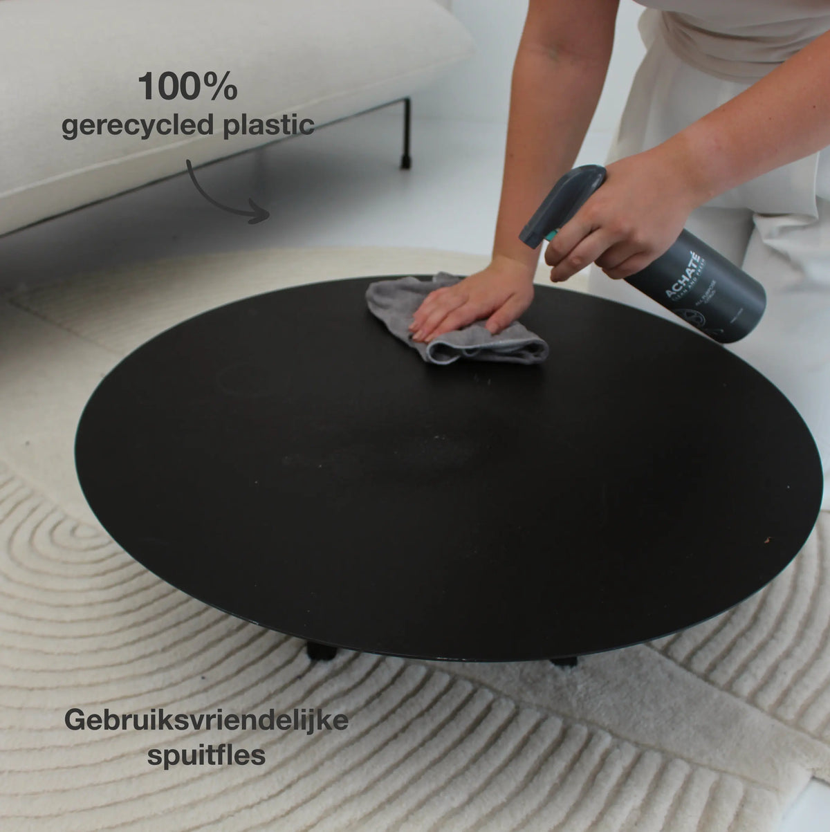 Person cleaning a black round table with a gray cloth and a spray bottle labeled for chemical-free cleaning, near a light-colored sofa and textured rug.