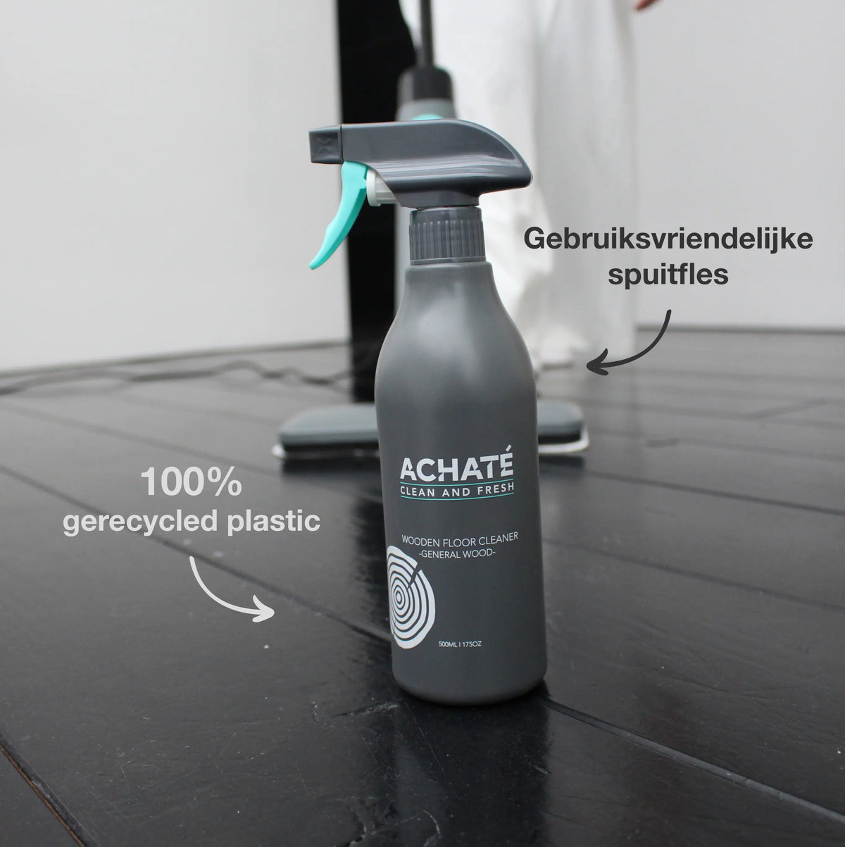 Dark gray spray bottle labeled Achaté Clean and Fresh wooden floor cleaner, positioned on a black wooden floor with a vacuum cleaner in the background, highlighting its user-friendly spray and 100% recycled plastic material.