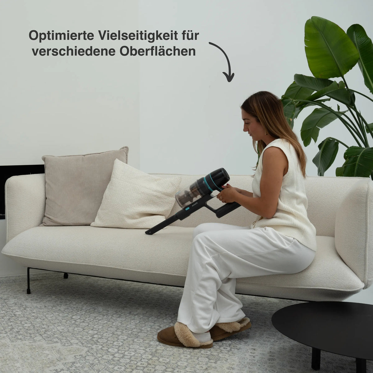 Woman using a cordless stick vacuum cleaner on a light-colored sofa in a modern living room with neutral decor and a large green plant nearby.