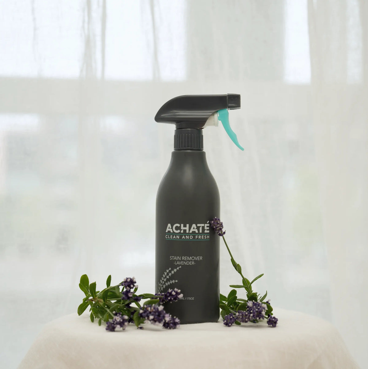 Black spray bottle labeled Achaté stain remover lavender with sprigs of lavender flowers around it on a white surface against a sheer curtain background