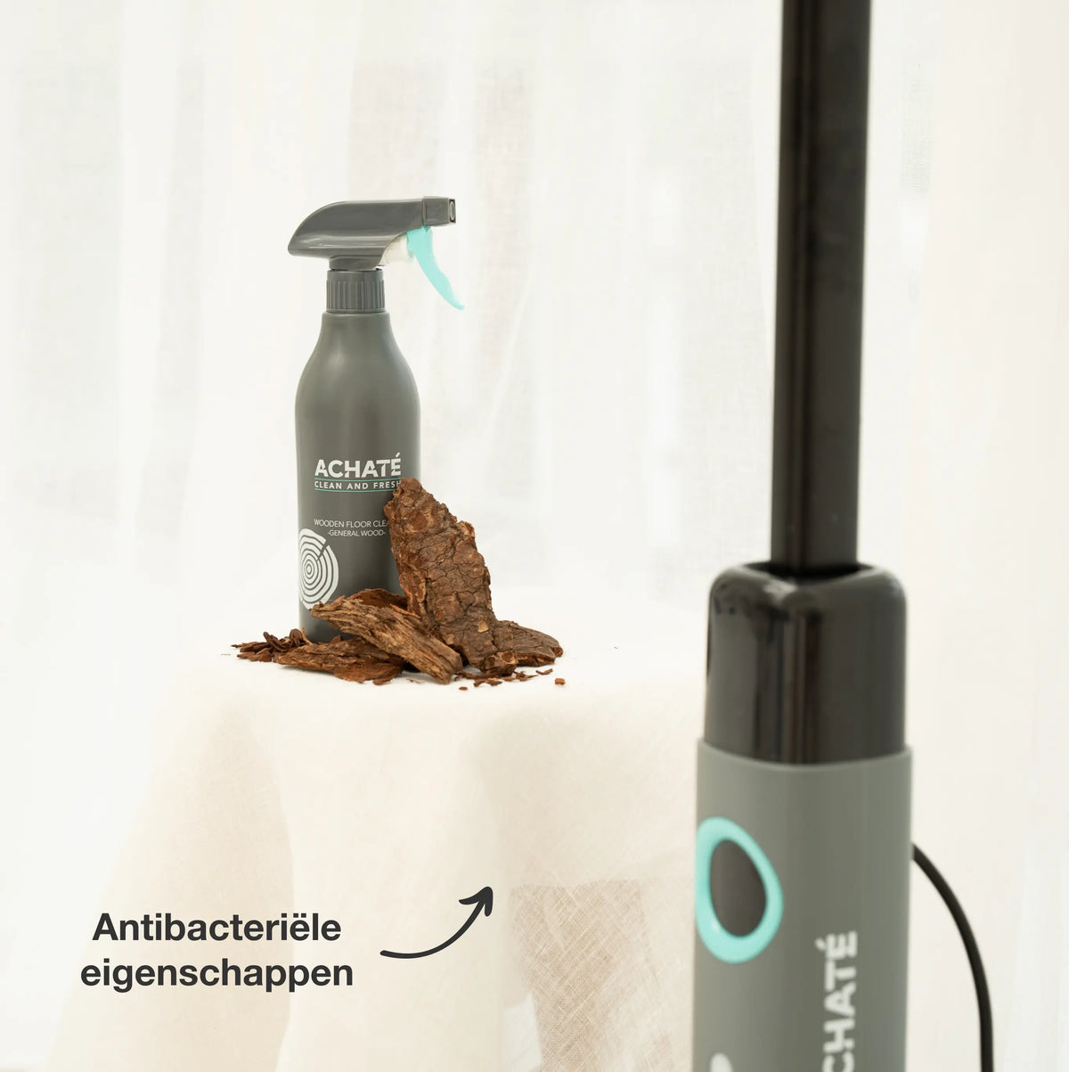 Spray bottle labeled Achaté with natural bark pieces nearby and part of a grey and black cleaning device with blue accent on a white fabric surface.