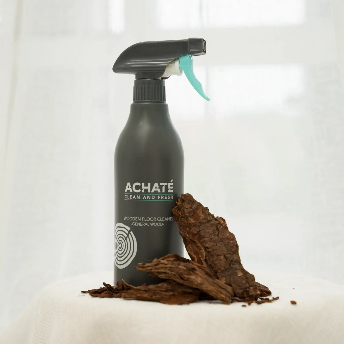 Black spray bottle labeled Achaté Clean and Fresh wooden floor cleaner next to pieces of natural wood bark on a light surface with a bright background.