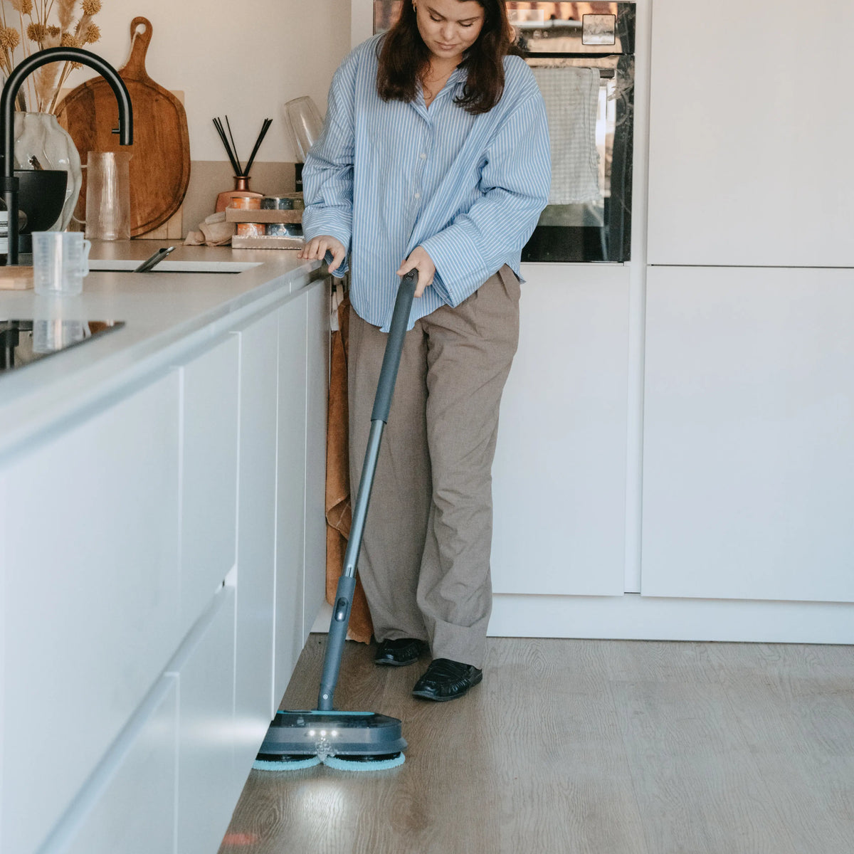 Woman using a cordless stick vacuum cleaner with two rotating cleaning pads to deep clean a light-colored kitchen floor next to white cabinets and a modern sink.