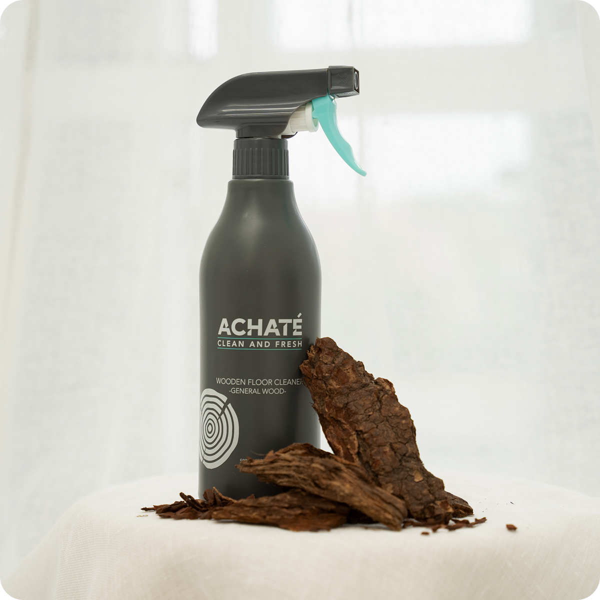 Matte black spray bottle labeled for wooden floor cleaning with dark brown bark pieces arranged beside it on a light fabric surface.