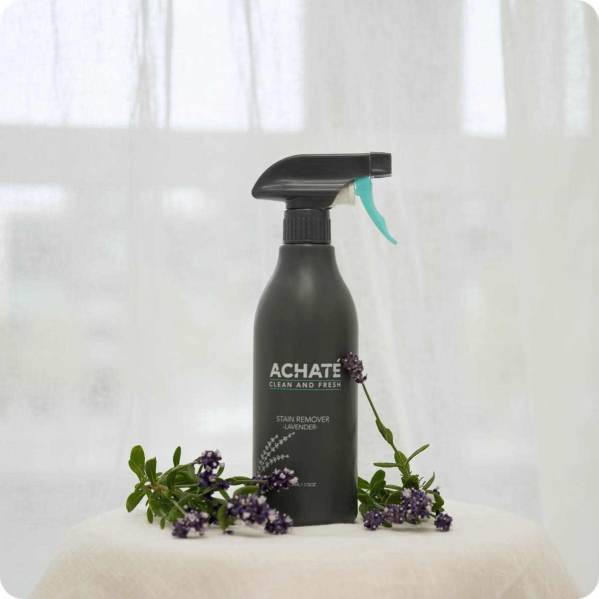 Black spray bottle labeled Achaté stain remover with lavender scent, surrounded by fresh lavender flowers on a white surface with sheer curtains in the background.