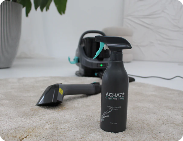 Black spray bottle labeled Achaté stain remover stands on a beige carpet with a handheld steam cleaner and its hose in the background on a light floor.