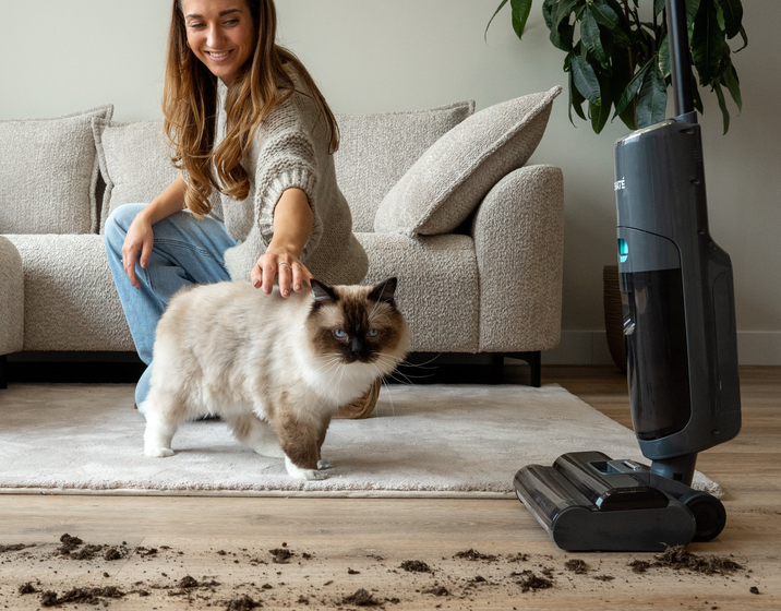 Achate Dweilstofzuiger V2 op een houten vloer met kruimels, vrouw en kat op de achtergrond