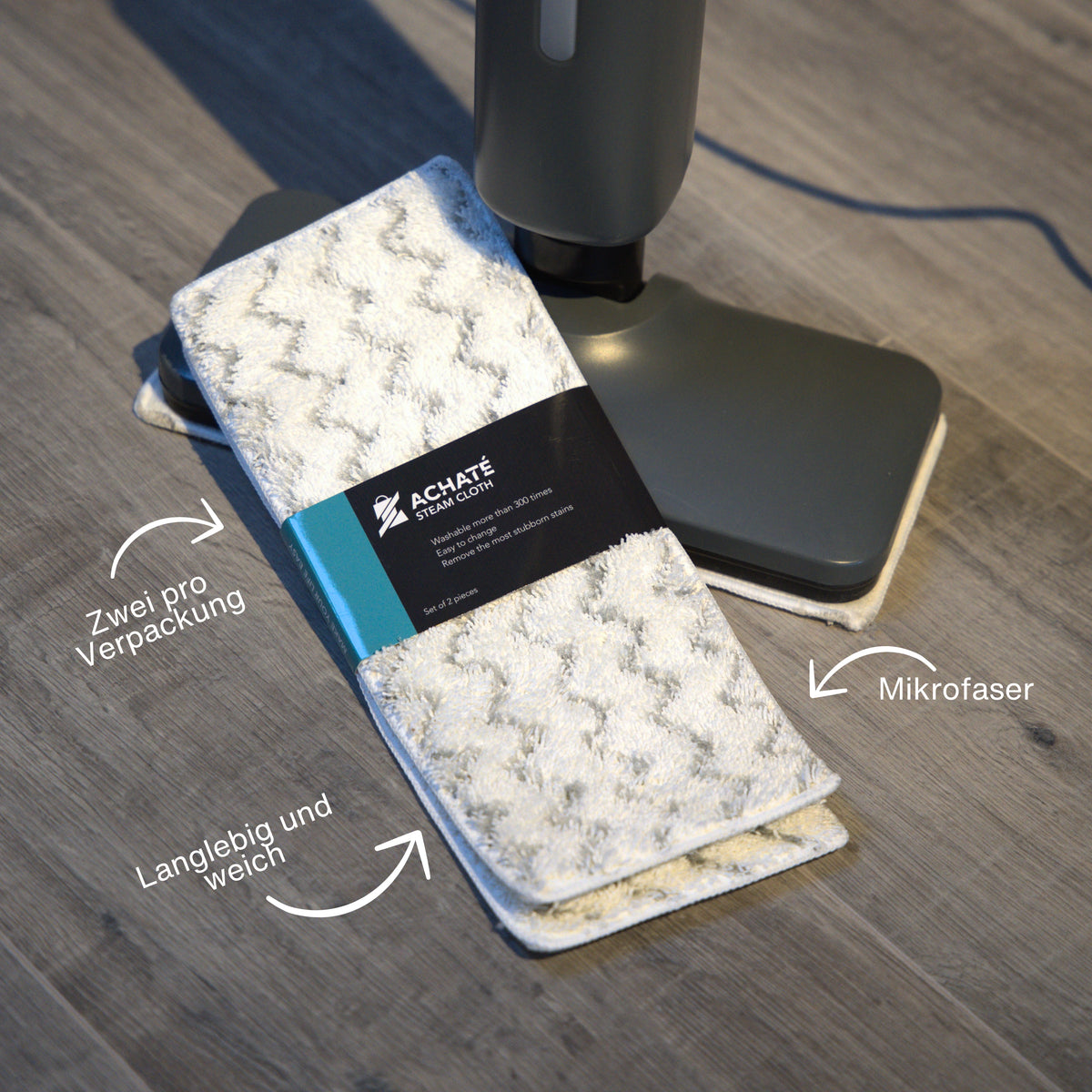 Set of two white microfiber steam cloths with zigzag pattern and a black wrap label, placed next to a dark gray steam mop head on a wooden floor.