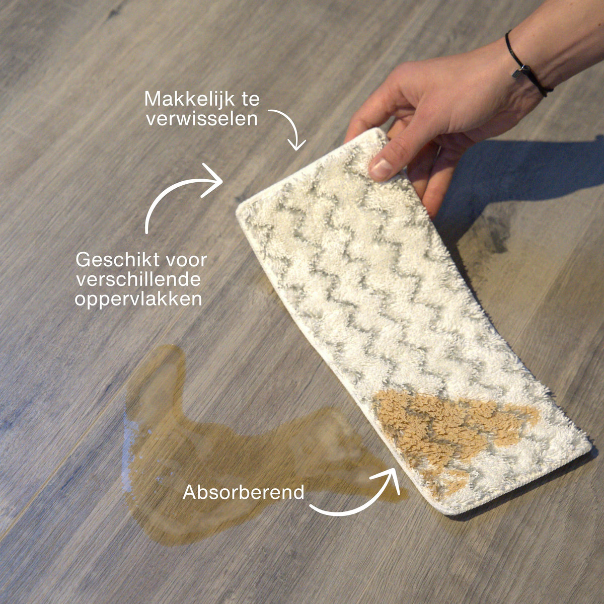 Hand holding a white and gray patterned microfiber cleaning pad absorbing a brown liquid spill on a wooden floor, showing easy-to-change and multi-surface cleaning features.