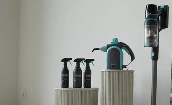 Three black spray bottles and a handheld steam cleaner displayed on two cylindrical pedestals next to a cordless stick vacuum cleaner mounted on the wall against a plain background.