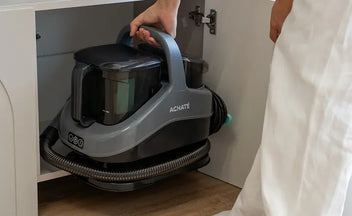 Person placing a grey Achaté handheld steam cleaner with hose and handle inside a lower kitchen cabinet, highlighting compact storage and cord management.