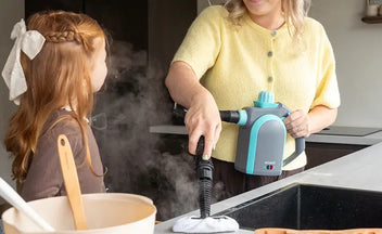 Woman using a handheld steam cleaner with high steam pressure to clean a kitchen countertop while a young girl watches nearby.